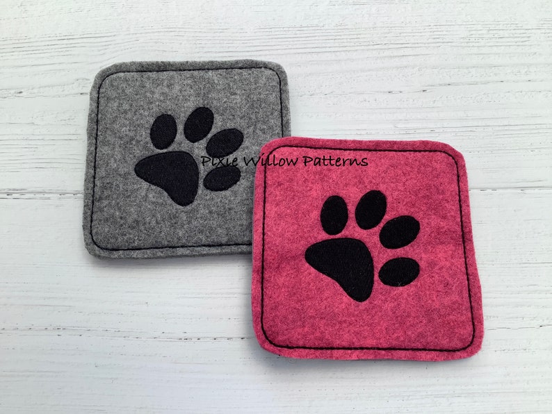 Puede incluir: Dos posavasos de fieltro con dise&ntilde;os de huellas de patas negras. Un posavasos es gris y el otro es rosa. Los posavasos est&aacute;n sobre una superficie de madera blanca. Pixie Willow Patterns.