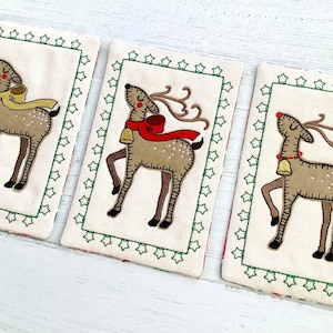 May include: Three embroidered Christmas ornaments featuring reindeer with red noses, antlers, and bells. Each ornament is framed with a green border of stars.
