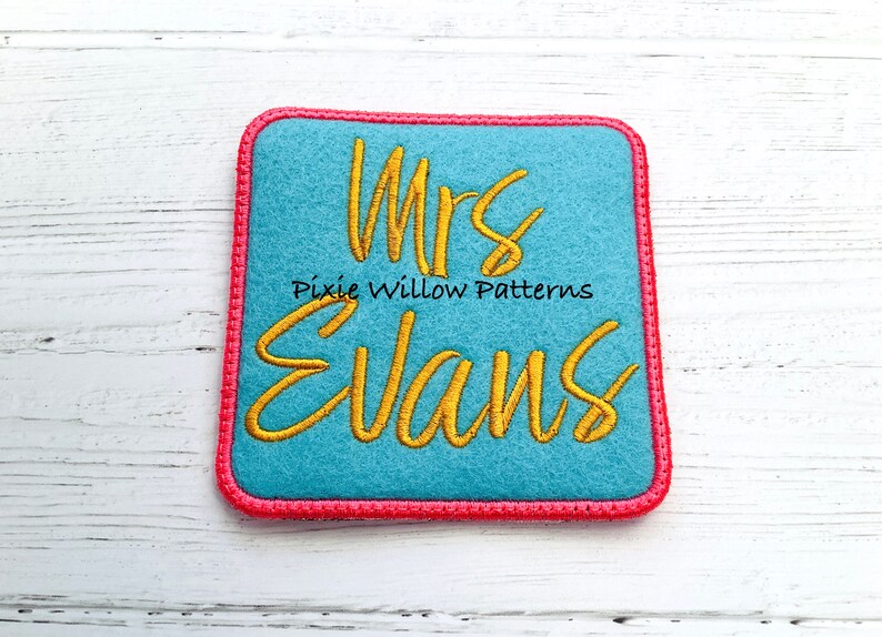 May include: A blue felt square with a pink border, featuring the embroidered text "Mrs. Evans" in gold thread. The text "Pixie Willow Patterns" is also embroidered in gold thread below the name.