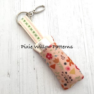 May include: A pink and white fabric keychain with a floral pattern and a small white tube inside. The keychain has a green and pink ribbon with a silver key ring. The text "Pixie Willow Patterns" is visible on the fabric.