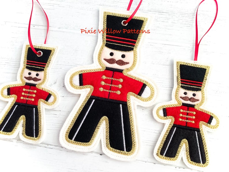May include: Three felt Christmas ornaments shaped like gingerbread men, each wearing a red and black uniform with gold trim. The ornaments are decorated with gold buttons and a black hat with gold trim. The largest ornament is approximately 6 inches tall, the middle ornament is approximately 4 inches tall, and the smallest ornament is approximately 3 inches tall.