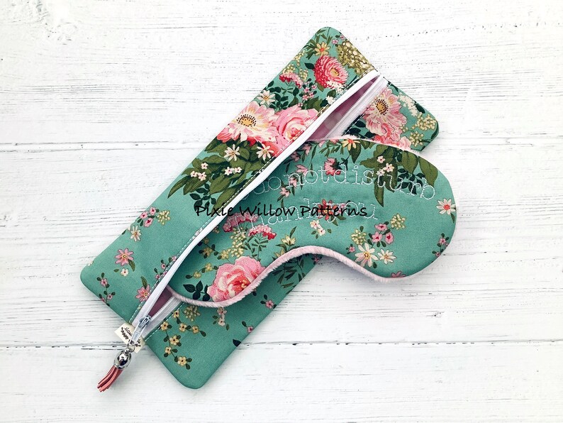 May include: A teal blue eye mask with a floral print and the text "do not disturb" embroidered on it. The eye mask is in a matching teal blue zippered pouch with a floral print.