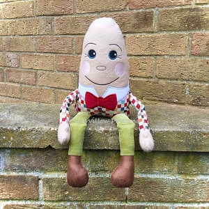Pode incluir: Um brinquedo de pelúcia em forma de ovo marrom com um rosto sorridente, usando uma gravata borboleta vermelha e uma camisa xadrez. O brinquedo está sentado em uma parede de tijolos.
