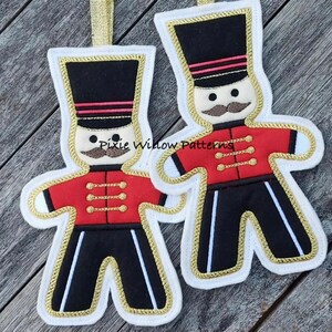 May include: Two felt Christmas ornaments shaped like gingerbread men, each wearing a red and black uniform with gold buttons and a black hat with a red stripe. The ornaments are decorated with gold stitching and have a white background.