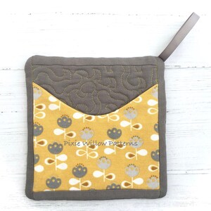 May include: A gray and yellow quilted pot holder with a floral pattern and a gray loop handle. The pot holder has the text "Pixie Willow Patterns" printed on it.