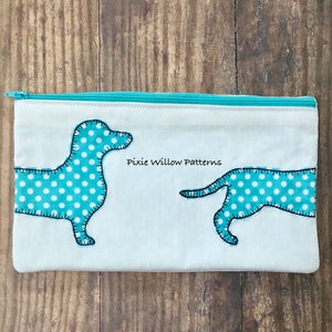 May include: A white zippered pouch with two dachshund dogs embroidered on it. The dogs are teal blue with white polka dots. The text "Pixie Willow Patterns" is embroidered on the pouch.