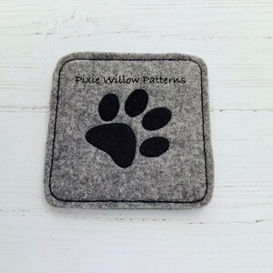 Puede incluir: Posavasos de fieltro gris con un dise&ntilde;o de huella de pata negra. El posavasos tiene el texto "Pixie Willow Patterns" bordado.