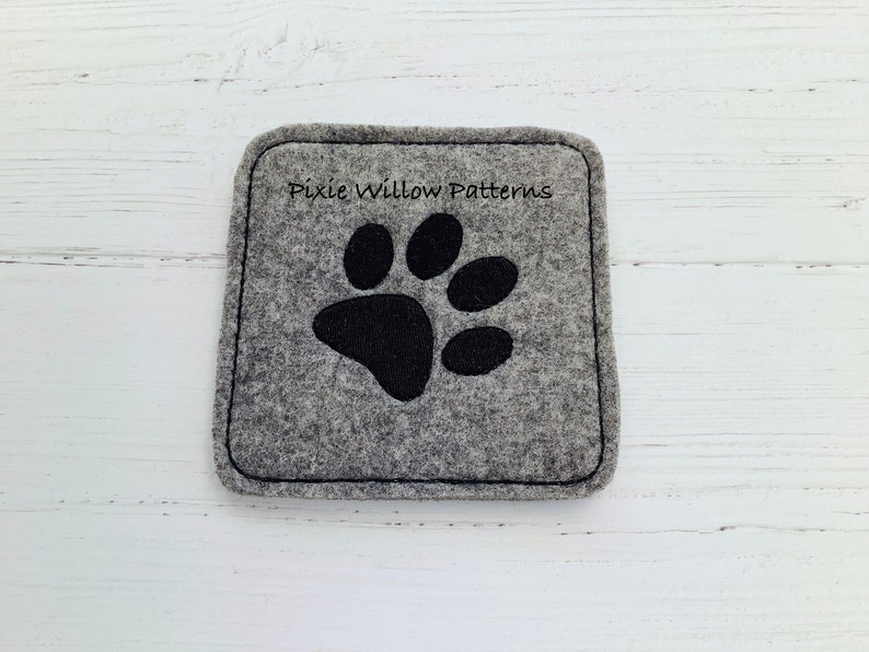 Puede incluir: Posavasos de fieltro gris con un dise&ntilde;o de huella de pata negra. El posavasos tiene el texto "Pixie Willow Patterns" bordado.