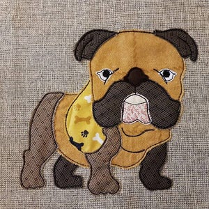 May include: A tan and brown bulldog applique with a yellow collar featuring bone and paw print designs. The dog has a patterned snout and ears, and a pink and white patterned mouth. The applique is on a gray textured background.