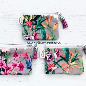 May include: Three small zippered pouches with floral patterns and pink tassels. The pouches are made of a soft, gray fabric with a white zipper. The floral patterns are different on each pouch. The pouches are labeled "Handmade with love".