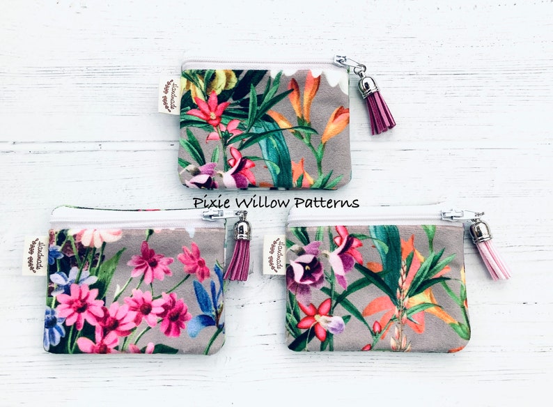 May include: Three small zippered pouches with floral patterns and pink tassels. The pouches are made of a soft, gray fabric with a white zipper. The floral patterns are different on each pouch. The pouches are labeled "Handmade with love".