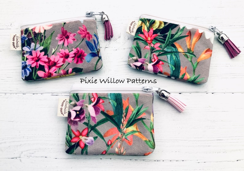 May include: Three small zippered pouches with floral patterns and tassels. The pouches are made of a soft, gray fabric with a white zipper. The tassels are pink, red, and white. The pouches are labeled "Pixie Willow Patterns".