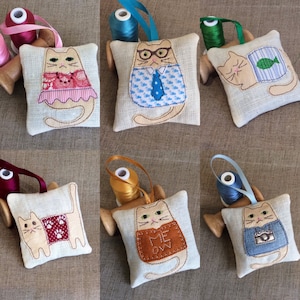 May include: Six handmade cat-shaped sachets with different embroidered designs. Each sachet is made of beige linen fabric and has a different embroidered design on the front. The designs include a cat wearing a red dress, a cat wearing a blue shirt and tie, a cat wearing a blue shirt with a camera, a cat wearing a brown vest with the words "Me Ow" on it, a cat wearing a red vest with paw prints, and a cat wearing a blue and white striped shirt with a fish.
