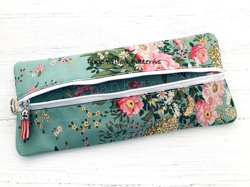 May include: A teal zippered pouch with a floral print and a white zipper. The pouch has a tassel pull and the text "Pixie Willow Patterns" is embroidered on the front.