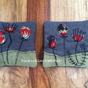 Puede incluir: Dos cuadrados de tela gris con flores bordadas en rojo, amarillo y negro. Los cuadrados de tela tienen un borde de tela verde con un diseño cosido. El texto "Pixie Willow Patterns" está bordado en la parte inferior de los cuadrados.