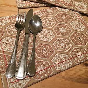 Pode incluir: Um conjunto de três talheres de prata, incluindo um garfo, uma faca e uma colher, são colocados em um descanso de mesa estampado em marrom e branco com costuras vermelhas ao redor das bordas.
