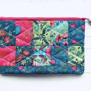 May include: A quilted zippered pouch with a patchwork design featuring floral and geometric patterns in shades of pink, green, and blue. The pouch has a zipper closure and a small tassel.