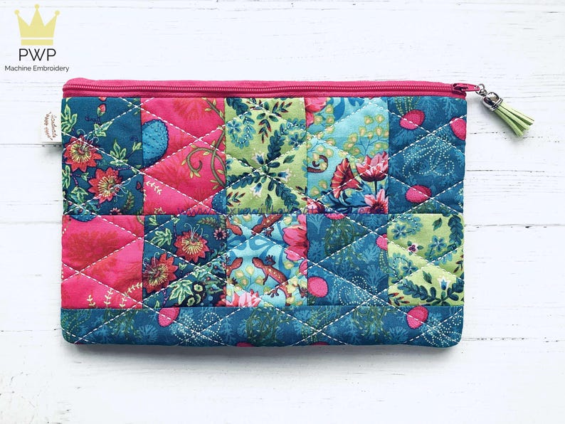 May include: A quilted zippered pouch with a patchwork design featuring floral and geometric patterns in shades of pink, green, and blue. The pouch has a zipper closure and a small tassel.