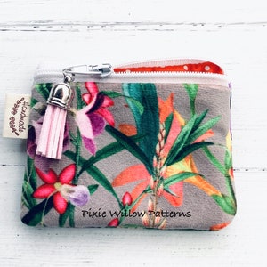 May include: A small gray velvet zipper pouch with a floral print featuring pink, orange, and green flowers. The pouch has a pink tassel attached to the zipper pull and a white label that says "Handmade with love". The pouch also has the text "Pixie Willow Patterns" printed on the bottom.