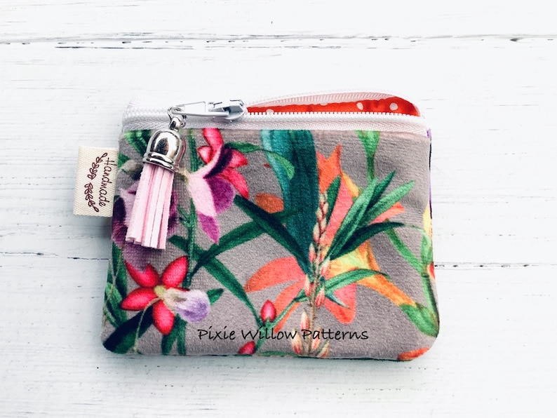May include: A small gray velvet zipper pouch with a floral print featuring pink, orange, and green flowers. The pouch has a pink tassel attached to the zipper pull and a white label that says "Handmade with love". The pouch also has the text "Pixie Willow Patterns" printed on the bottom.