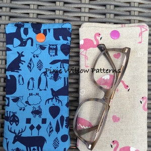 May include: Two eyeglass cases. One is light blue with a dark blue animal pattern and an orange button closure. The other is beige with a pink flamingo pattern and a pink button closure. Eyeglasses rest on top of the flamingo case.
