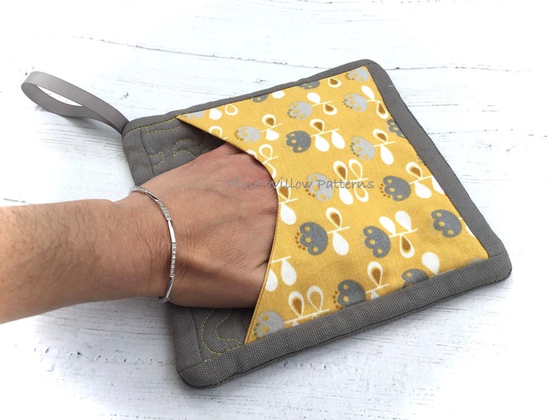May include: A gray and yellow pot holder with a floral pattern and a gray loop handle. The pot holder has a pocket for a hand.