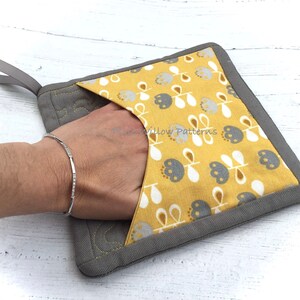 May include: A gray and yellow pot holder with a floral pattern and a gray loop handle. The pot holder has a pocket for a hand.