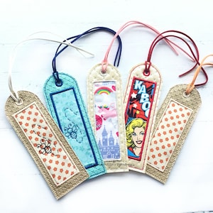 May include: Five colourful fabric bookmarks with different designs. The bookmarks are made of a beige fabric with a quilted texture and have a loop at the top for hanging. The designs include a bee, a castle, a comic book character, and two different floral patterns.