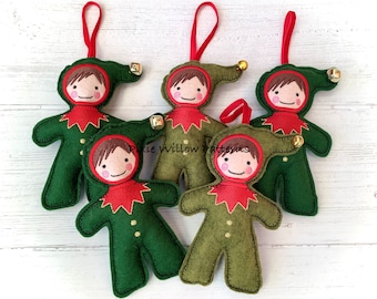 May include: Five felt Christmas ornaments shaped like elves with green bodies, red hats, and brown hair. Each elf has a gold bell on its hat and a gold star on its chest. The ornaments are hanging from red ribbons.