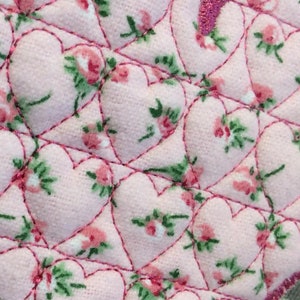 Może przedstawiać: Różowy materiał z pikowanym wzorem małych serc. Serca są obwiedzione różową nicią, a materiał ma kwiatowy nadruk.