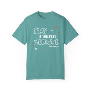 May include: A teal blue t-shirt with the text "PLAY IS THE BEST MEDICINE" in white with a white star and orange star design. The text "Child Family Life" is printed in smaller white text below the main text.