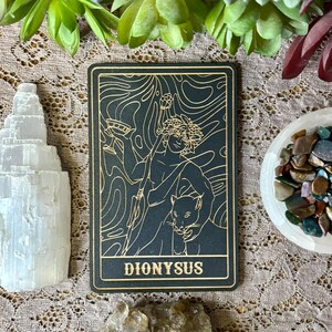Op de afbeelding: Een zwarte en gouden tarotkaart met een illustratie van Dionysus met een panter. De kaart heeft een gouden rand en het woord "DIONYSUS" onderaan. Kristallen en groen omringen de kaart.