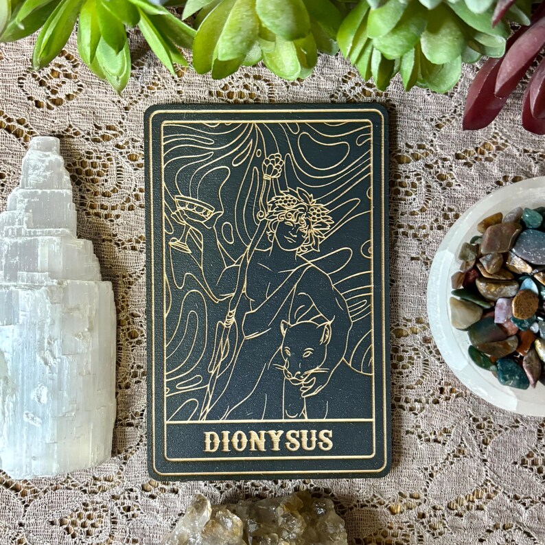 Op de afbeelding: Een zwarte en gouden tarotkaart met een illustratie van Dionysus met een panter. De kaart heeft een gouden rand en het woord "DIONYSUS" onderaan. Kristallen en groen omringen de kaart.