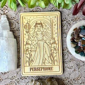 Pode incluir: Um cart&atilde;o de madeira com um desenho de linha de Pers&eacute;fone, a deusa grega do submundo, sentada em um trono. Ela segura uma rom&atilde; em sua m&atilde;o. O cart&atilde;o &eacute; rotulado como "PERSEPHONE".