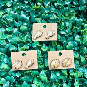 May include: Wooden stud earrings in the shape of chicken drumsticks and wings. The earrings are attached to small, rectangular, brown cardboard earring cards. The earrings have a gold glittery finish and are set against a green leafy background.