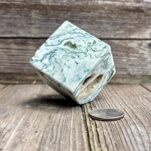May include: A cube-shaped stone with a white and green marbled pattern featuring dark green moss-like inclusions and a small natural cavity. A silver-colored United States quarter coin is placed next to the stone for scale on a wooden surface.