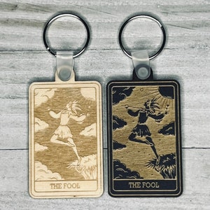 May include: Two wooden keychains featuring the Major Arcana tarot card "The Fool". One keychain is light brown with a black design, and the other is dark brown with a light brown design.