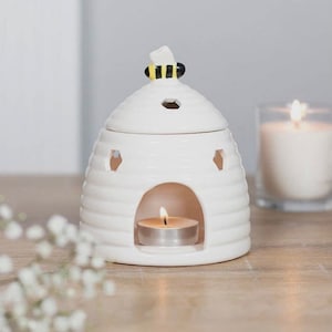 May include: A white ceramic essential oil diffuser shaped like a beehive, with a small bee figurine on top. The diffuser has a small opening in the front for a tea light candle, which is lit. The background includes a lit candle in a glass and some white flowers.