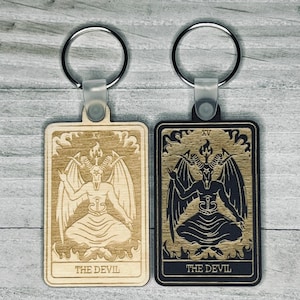 May include: Two wooden keychains featuring the "The Devil" card from a tarot deck. One keychain is light wood with a dark brown image, the other is dark wood with a light brown image.
