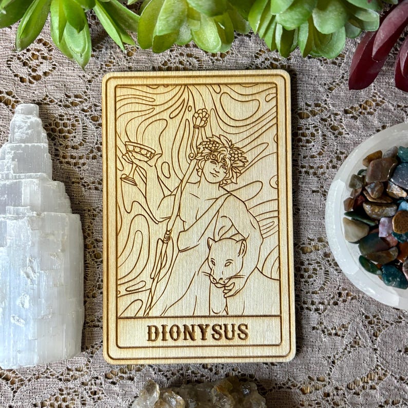 Op de afbeelding: Een houten tarotkaart met een gegraveerde afbeelding van Dionysus, met een beker en staf, met een panter. Het woord "DIONYSUS" is onderaan gegraveerd. De kaart is omgeven door andere decoratieve items.