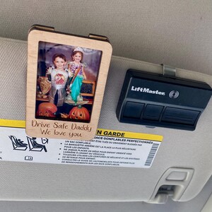 May include: A wooden photo frame with a picture of two children dressed in Halloween costumes. The frame is attached to a car's sun visor. The text "Drive Safe Daddy We love you." is written on the frame.