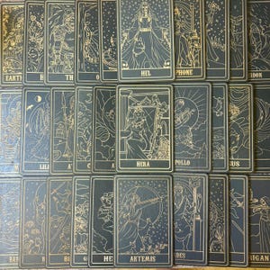 Op de afbeelding: Een verzameling tarotkaarten met gouden lijntekeningen op een donkere achtergrond. Elke kaart toont een ander figuur of symbool, met namen als "HEL", "HERA" en "ARTEMIS" erop gedrukt. De kaarten zijn in een raster gerangschikt.