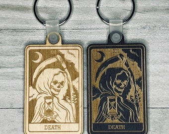May include: Two wooden keychains featuring the Death tarot card. One keychain is light wood with a dark brown image, the other is dark wood with a light brown image. Both keychains feature the Death card number, XIII, and the word "DEATH" at the bottom.
