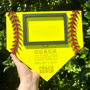 May include: A yellow wooden softball home plate with a black frame picture holder. The home plate has red stitching and a gold inscription that reads: "A good coach can change a game. A great coach can change a life. Coach Devin."