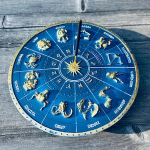 May include: A blue and gold incense holder featuring a zodiac wheel design. The holder has raised gold zodiac symbols and text, with an incense stick in the center. The background is a weathered wooden surface.
