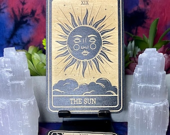 May include: A wooden tarot card with a black background and gold accents. The card depicts a sun with a face and rays. The card number XIX and the words "THE SUN" are printed on the card.