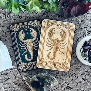 May include: Two wooden engraved cards with a scorpion design. The card on the left has a dark green background and the card on the right has a light wood background. Both cards have the word "SCORPIO" written on them.