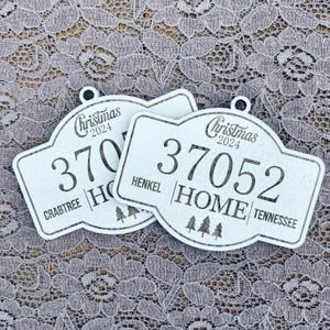 May include: Two white wooden Christmas ornaments with the text "Christmas 2024" and a house address. The ornaments feature a stylized house design with a number, street name, and state. The first ornament reads "370 CRABTREE | HOME" and the second ornament reads "37052 HENKEL | HOME | TENNESSEE".
