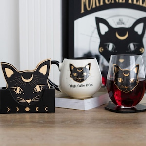 May include: A black cat-themed home decor set. Includes a black coaster set, a white mug with "Magic, Coffee & Cats" text, and a stemless wine glass, all featuring a black cat design with gold accents. A black and gold framed print is in the background.