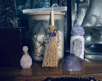May include: A display of spiritual items on a wooden surface. Features a candle in a glass jar with a pentagram design, a small broom with a pentacle charm, a rose quartz perfume bottle, and an amethyst crystal.